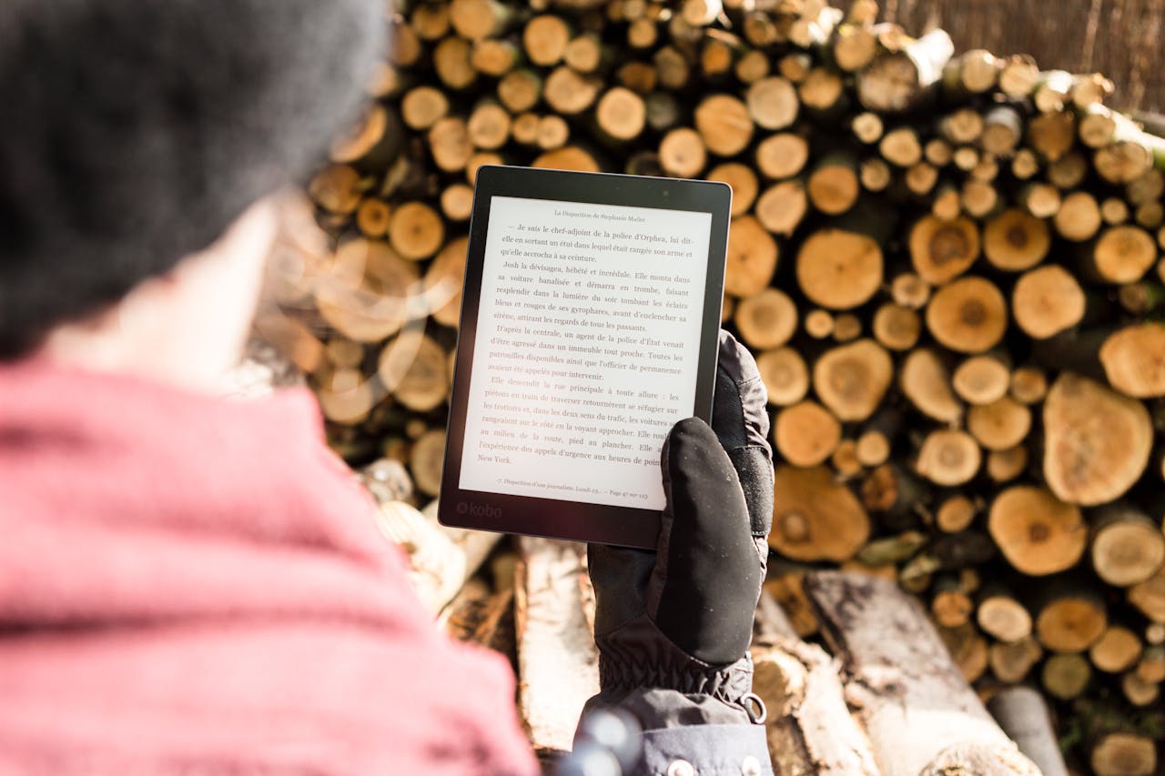 Person in warm clothing reads an e-book outdoors near a pile of firewood.